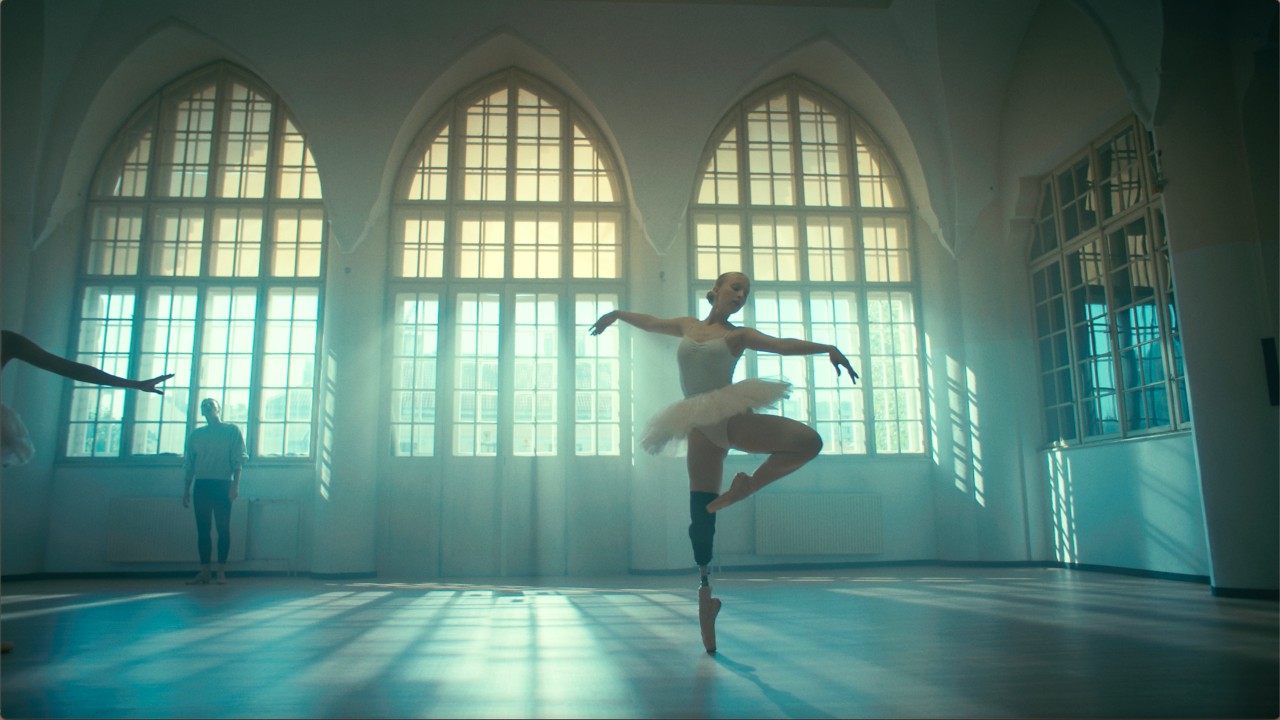O balerină cu un picior protetic stă nemișcată în poziție într-un studio tip loft în care lumina se filtrează prin ferestrele imense din spatele ei.